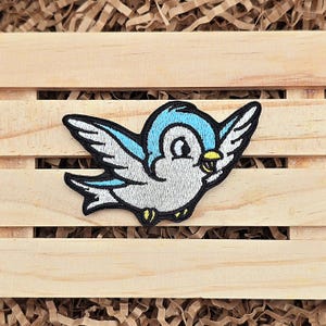 May include: A blue and white embroidered patch of a cartoon bird with a yellow beak. The bird is flying with its wings spread out.