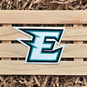May include: Embroidered patch featuring the letter "E" in white, outlined with black and teal. The patch has a stylized, angular design. The patch is placed on a wooden crate with brown paper filler.