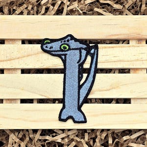 May include: A light blue dinosaur patch with a black outline and green eyes. The patch is embroidered and has a textured appearance. The patch is on a wooden surface.