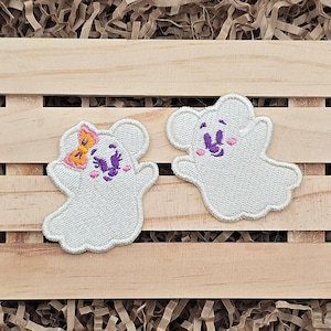May include: Two white ghost-shaped embroidered patches. The left patch features a yellow and pink bow, purple eyes, and pink cheeks. The right patch features purple eyes and pink cheeks. Both patches have a white stitched border.