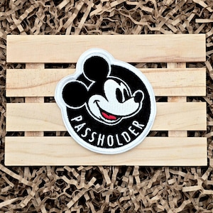May include: A circular patch featuring Mickey Mouse with the text "PASSHOLDER" in white. The patch has a black background with a white border. The image is set on a wooden pallet.