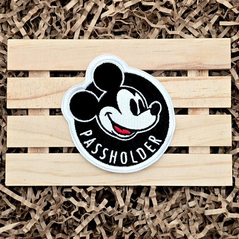 Op de afbeelding: Een ronde patch met Mickey Mouse met de tekst "PASSHOLDER" in het wit. De patch heeft een zwarte achtergrond met een witte rand. De afbeelding is geplaatst op een houten pallet.