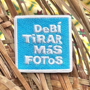 May include: Square embroidered patch with the text "Debí Tirar Más Fotos" in white on a light blue background, framed by a white border. The patch is set against a woven background.