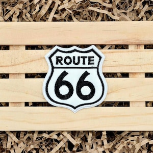 May include: A white Route 66 patch with black border and text. The patch features the words "ROUTE" above the number "66" in a shield shape. The patch is on a wooden crate with brown paper filler.