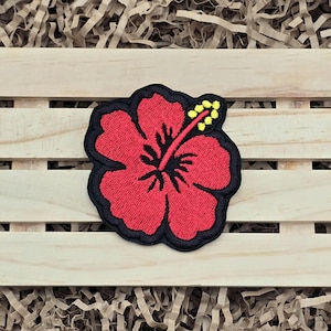May include: Embroidered red hibiscus flower patch with a black border and yellow stamens. The patch is a vibrant representation of a tropical flower, perfect for adding a touch of the tropics to clothing or accessories.