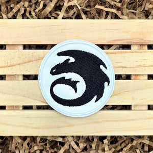 May include: A round, embroidered patch featuring a black dragon design on a light blue background. The patch is set against a wooden crate filled with brown paper. The dragon design is a stylized silhouette.