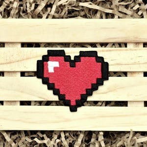 May include: A pixelated heart patch with a red center, black border, and white accent. The patch is displayed on a light wooden surface with shredded paper. This patch is perfect for adding to clothing or accessories.