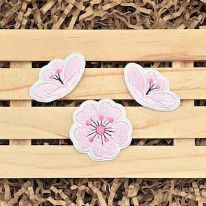 May include: Three embroidered patches featuring cherry blossom designs. Two patches are butterfly-shaped, and one is a circular flower. The patches are light pink with white borders and black and pink details.