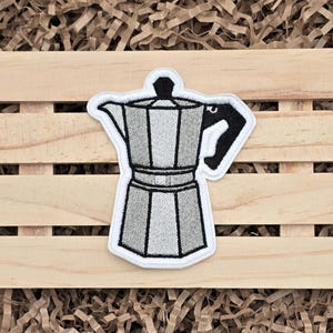 May include: Embroidered patch featuring a gray and black coffee maker design with a white border. The patch is a detailed illustration of a stovetop espresso maker, perfect for coffee lovers. The patch is set against a wooden background.
