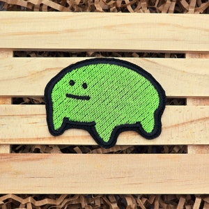 May include: A green embroidered patch with a black outline, featuring a simple cartoon character with a small mouth and two dots for eyes. The patch is on a wooden surface with brown paper filler.