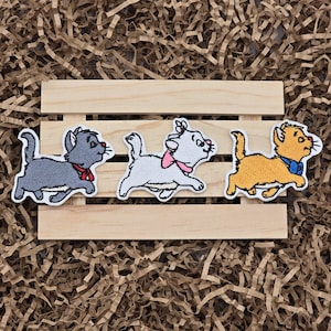 May include: Three embroidered iron-on patches featuring characters from the Disney movie The Aristocats. The patches are of a gray cat, a white cat, and an orange cat. The patches are on a wooden background.