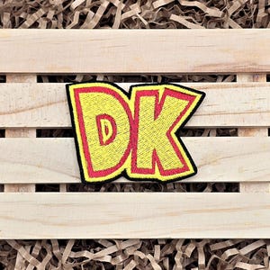 May include: Embroidered patch featuring the letters "DK" in yellow with a red outline and black border. The patch is placed on a light-colored wooden crate with shredded paper in the background.