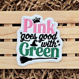 May include: Embroidered patch with the text "Pink goes good with Green". The patch features pink and green lettering, a black witch hat, and a black broom. The patch has a white border and a black crown.