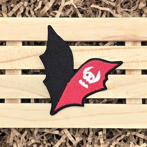 May include: Embroidered patch in the shape of a bat wing, with a black wing on the left and a red wing on the right. The red wing features the number "18" in white. The patch is placed on a wooden surface.