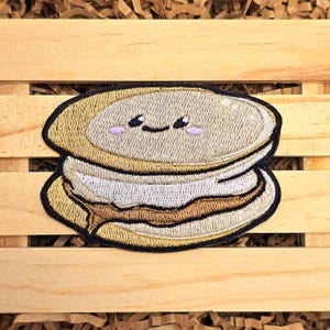 May include: A cute embroidered patch of a smiling, layered dessert with brown, white, and beige frosting.
