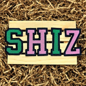 May include: Embroidered patches spelling out the word "SHIZ" in a collegiate font. The letters are green and pink with a black border, set on a wooden pallet, surrounded by brown paper filler.