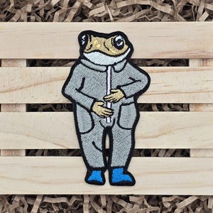 May include: Embroidered patch featuring a frog character in a gray jumpsuit with blue boots. The frog has a tan head, white collar, and is holding a white object. The patch is on a wooden background.