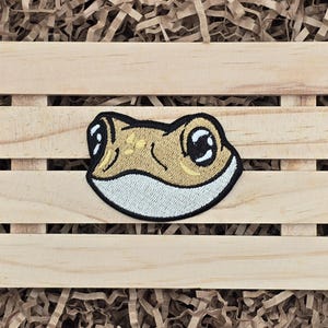 May include: Embroidered patch of a frog's face with large, black eyes and a light brown body. The frog is smiling with its mouth closed.