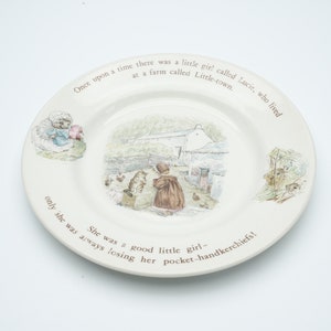 Peut inclure: Une assiette en céramique blanche avec une bordure brune et une illustration colorée d'une fille et d'un chat marchant sur un chemin vers une maison. L'assiette a du texte autour de la bordure qui dit "Il était une fois une petite fille nommée Lucie, qui vivait dans une ferme appelée Little-town. Elle était une bonne petite fille - seulement elle perdait toujours ses mouchoirs!"