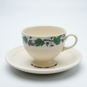 May include: A white teacup and saucer with a green vine pattern. The cup has a fluted design and a handle.