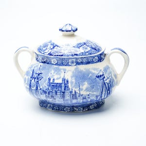 Può includere: Una zuccheriera in ceramica bianca con un motivo blu a trasferimento raffigurante un castello, velieri e bordi floreali. Il contenitore è dotato di due manici laterali e un coperchio coordinato con un piccolo pomello come impugnatura.