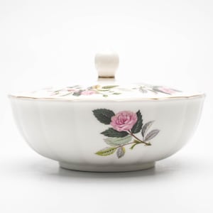 Op de afbeelding: Witte porseleinen kom met een deksel, met een ontwerp van een roze roos met groene bladeren. De kom heeft een gouden rand en een gegolfde rand.