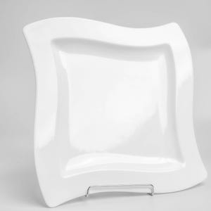 Op de afbeelding: Een witte, vierkante keramische plaat met een golvende rand. De plaat wordt op een metalen standaard gepresenteerd tegen een effen witte achtergrond. Het glanzende oppervlak van de plaat reflecteert licht.