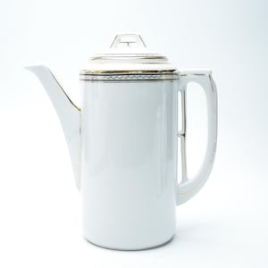 Puede incluir: Una cafetera de cerámica blanca con un cuerpo alto y cilíndrico y un pico largo. La cafetera tiene tapa y asa. Los adornos dorados acentúan el borde, la tapa y el asa. La cafetera es probablemente vintage o antigua.