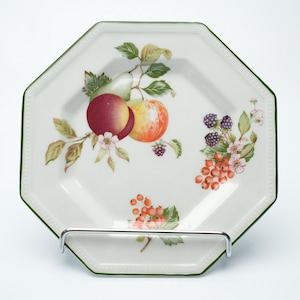 Puede incluir: Un plato octogonal blanco con un borde verde y un diseño de frutas. El plato presenta ilustraciones de manzanas, bayas y grosellas. El plato se muestra en un soporte de metal plateado.
