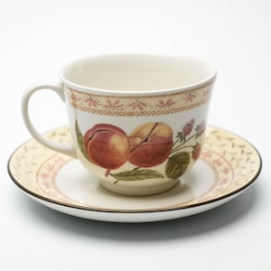 Puede incluir: Una taza de té y un platillo blancos con un diseño de melocotón y flores. La taza tiene un asa y el platillo tiene un borde festoneado.