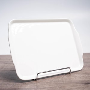 Puede incluir: Una bandeja de servir de cerámica blanca con un borde ligeramente redondeado y un soporte de metal. La bandeja está vacía y lista para ser utilizada.