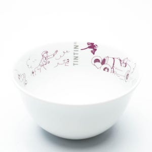 May include: White ceramic bowl featuring a maroon illustration of Tintin and Snowy. The bowl has a rounded shape and a smooth surface. The word "TINTIN" is printed on the side of the bowl.