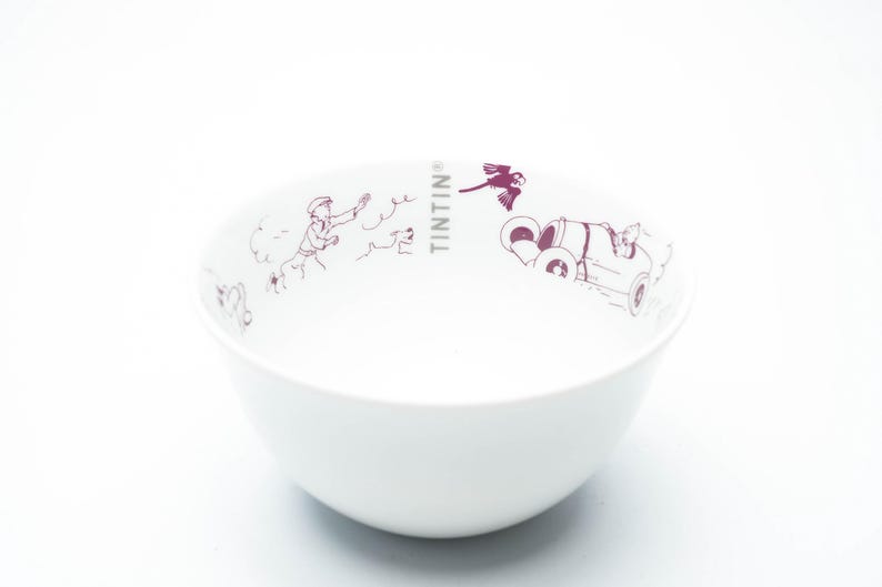 May include: White ceramic bowl featuring a maroon illustration of Tintin and Snowy. The bowl has a rounded shape and a smooth surface. The word "TINTIN" is printed on the side of the bowl.