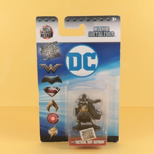 May include: A plastic blister pack containing a die-cast metal figurine of Batman in a tactical suit. The packaging is blue and features the DC Comics logo and the words "Justice League" and "Nano Metalfigs".