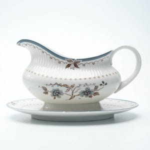 Könnte beinhalten: Weißes Keramik-Sauciere mit passender Untertasse. Das Gefäß hat einen blauen Rand und ein Blumenmuster in Blau und Braun. Es ist für das Servieren von Saucen und Soßen konzipiert, ideal für das Abendessen.