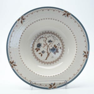 Könnte beinhalten: Eine weiße Keramikschale mit blauem Rand und floralem Design. Die Schale hat ein zentrales Blumenmotiv mit braunen und blauen Akzenten. Der Rand ist mit einem sich wiederholenden Muster aus kleinen braunen Blättern und Punkten verziert.