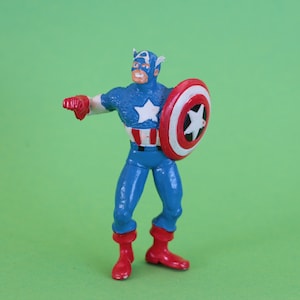 K&ouml;nnte beinhalten: Eine Plastik-Actionfigur von Captain America, der einen blauen und roten Anzug mit einem wei&szlig;en Stern auf der Brust tr&auml;gt. Er h&auml;lt einen rot-wei&szlig;-blauen Schild in seiner rechten Hand.
