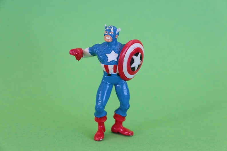 K&ouml;nnte beinhalten: Eine Plastik-Actionfigur von Captain America, der einen blauen und roten Anzug mit einem wei&szlig;en Stern auf der Brust tr&auml;gt. Er h&auml;lt einen rot-wei&szlig;-blauen Schild in seiner rechten Hand.
