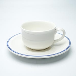 May include: A white ceramic teacup and matching saucer set. The teacup features a smooth, rounded bowl with a curved handle. The saucer has a circular design with a thin, distinct blue line accenting the outer rim, providing a clean look.