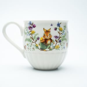 Puede incluir: Taza de cerámica blanca con asa curva, con una ilustración detallada de un conejo marrón sosteniendo un huevo, rodeado de flores y follaje coloridos. La taza tiene una base acanalada y un acabado liso y brillante.