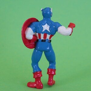K&ouml;nnte beinhalten: Eine Plastik-Actionfigur von Captain America, der sein ikonisches rot-wei&szlig;-blaues Kost&uuml;m tr&auml;gt. Er h&auml;lt einen rot-wei&szlig;-blauen Schild in seiner linken Hand und sein rechter Arm ist erhoben. Die Figur steht auf einem gr&uuml;nen Hintergrund.