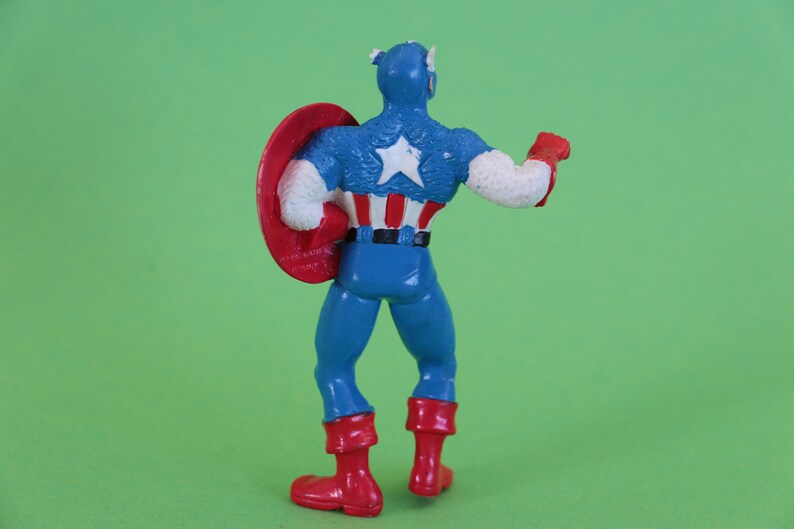 K&ouml;nnte beinhalten: Eine Plastik-Actionfigur von Captain America, der sein ikonisches rot-wei&szlig;-blaues Kost&uuml;m tr&auml;gt. Er h&auml;lt einen rot-wei&szlig;-blauen Schild in seiner linken Hand und sein rechter Arm ist erhoben. Die Figur steht auf einem gr&uuml;nen Hintergrund.
