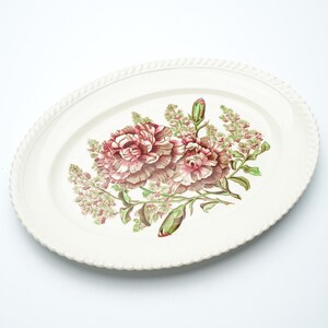Könnte beinhalten: Ovale Servierplatte mit cremefarbenem Hintergrund und floralem Design. Die Blumen sind rosa und weiß, mit grünen Blättern und Stielen. Der Rand der Platte hat eine seilartige Textur.
