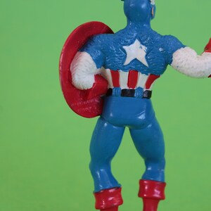 K&ouml;nnte beinhalten: Eine Plastik-Actionfigur von Captain America, der ein blaues und rotes Kost&uuml;m mit einem wei&szlig;en Stern auf der Brust tr&auml;gt. Er h&auml;lt einen rot-wei&szlig;-blauen Schild in seiner linken Hand.