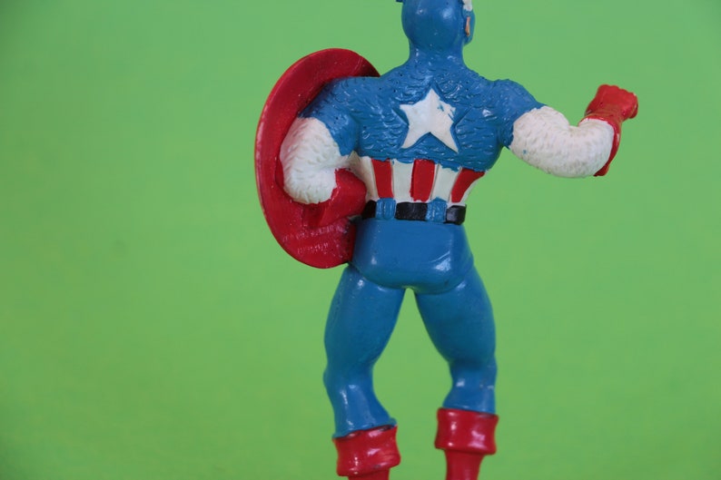 K&ouml;nnte beinhalten: Eine Plastik-Actionfigur von Captain America, der ein blaues und rotes Kost&uuml;m mit einem wei&szlig;en Stern auf der Brust tr&auml;gt. Er h&auml;lt einen rot-wei&szlig;-blauen Schild in seiner linken Hand.