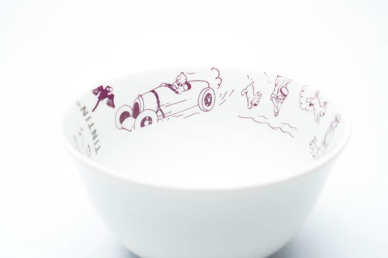 May include: White ceramic bowl with a maroon illustration of a car, bird, and characters from the Tintin comic series. The bowl has a simple, rounded shape and is suitable for serving cereal or soup.