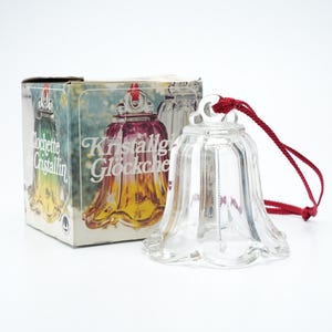 May include: A clear glass bell with a red string, next to a box with the words "Kristallig Glockchen" and "Clochette Cristallin". The bell has a scalloped edge and a small loop at the top for the string. The box has images of the bell in different colours.
