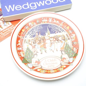 Op de afbeelding: Een decoratieve plaat met een rood-witte rand met een winterse scène met figuren, bomen en gebouwen. De plaat is gemarkeerd met "A Child's Christmas 1979". Een blauwe doos met "Wedgwood" en een boekje zijn ook aanwezig.