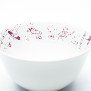 May include: White ceramic bowl with a maroon illustration of characters from the Tintin comic series. The bowl features a rounded shape and a smooth, glossy finish. The word "TINTIN" is printed on the side of the bowl.