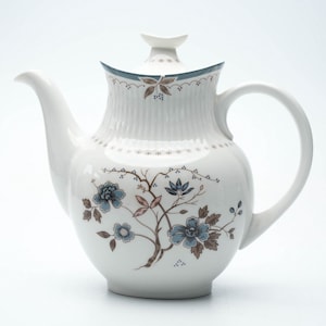 Può includere: Teiera in ceramica bianca con beccuccio e manico curvi. La teiera presenta un motivo floreale con fiori e rami blu e marroni. Il coperchio ha un piccolo pomello e un bordo decorativo.